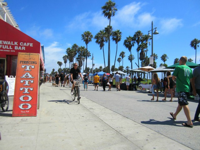 Venice boardwalk. A sign advertises tattoos and a bike rides toward the camera